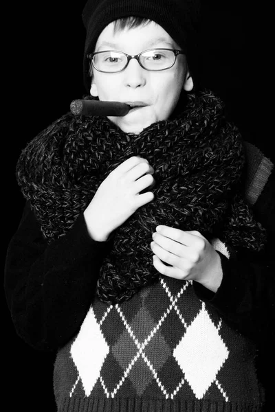 Ragazzo nerd in occhiali con sigaretta ragazzo sorridente o simpatico bambino nerd in occhiali, cappello e sciarpa lavorata a maglia alla moda su sfondo nero fumare sigaro . — Foto Stock