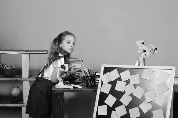 Menina coloca livros na mesa com microscópio e quadro-negro — Fotografia de Stock