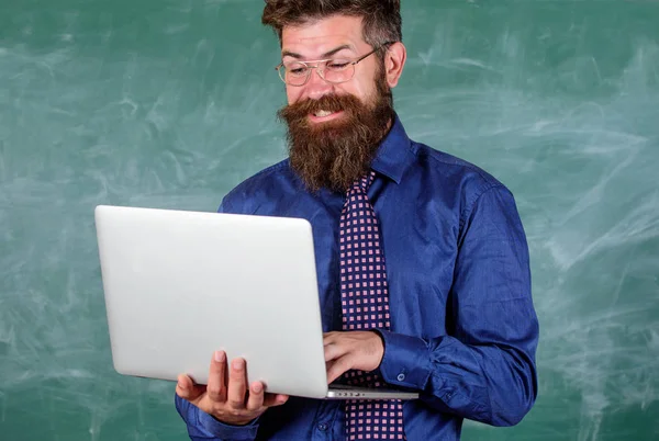 Surfing internet. Teacher bearded man with modern laptop surfing internet chalkboard background. Distance education concept. Hipster teacher wear eyeglasses and necktie holds laptop surfing internet