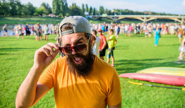 Празднование городского события. Человек смотрит через солнечные очки солнечный день на открытом воздухе. Хипстер в кепке посещает фестиваль пикников. Человек бородатый хипстер перед толпой людей зеленый фон поля
