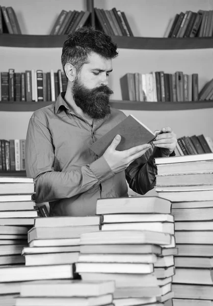 Homem em ocupado livro de leitura de rosto pensativo, prateleiras de livros no fundo. Professor ou estudante com barba estudando na biblioteca entre pilhas de livros. Conceito de educação e ciência — Fotografia de Stock
