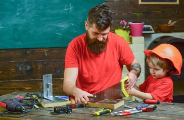 Masculine duties concept. Boy, child busy in protective helmet learning to use handsaw with dad. Father, parent with beard teaching little son to sawing with sharp handsaw, carpenter crafts clipart
