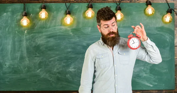 El profesor de anteojos tiene despertador. Concepto de horario escolar. Hombre con barba y bigote en la cara interesada de pie en el aula. hipster barbudo sostiene reloj, pizarra en el fondo, espacio de copia —  Fotos de Stock