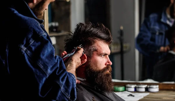 Il barbiere con tagliacapelli lavora sull'acconciatura per lo sfondo del barbiere. Barbiere che acconciano capelli di brutale cliente barbuto con clipper. Un cliente hipster che si taglia i capelli. Concetto di stile di vita hipster — Foto Stock