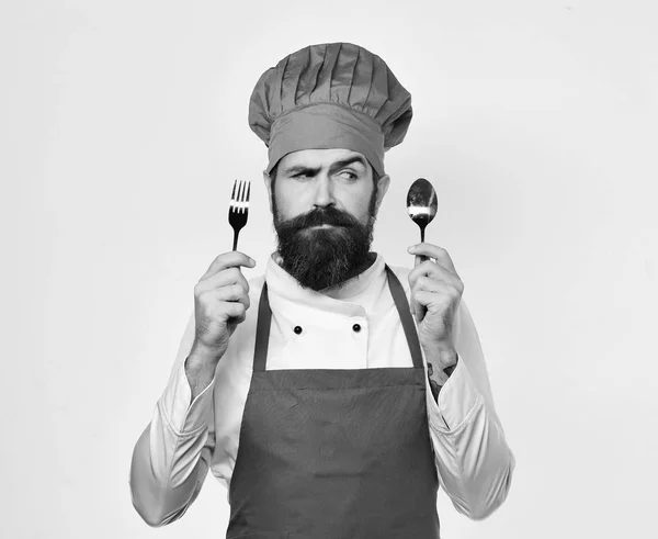 Cook with confused face in burgundy uniform shows cutlery — Stock Photo, Image