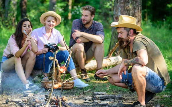 Friends group tourist relaxing near bonfire. Tourists having snack time with roasted over fire food. Hipster roasting sausage while friends sit on log sharing impression and watching photos on camera — Stock Photo, Image