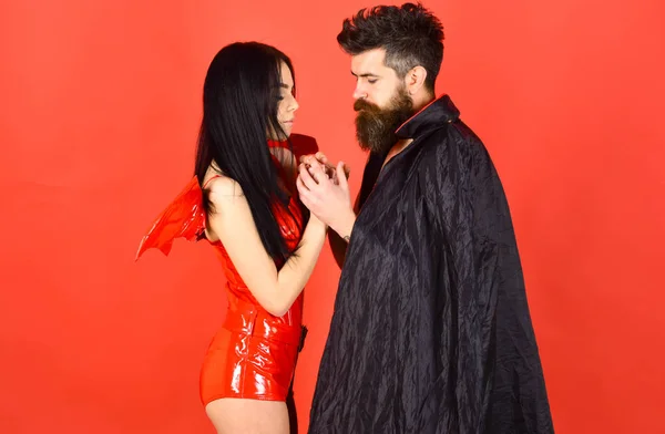 Couple on pensive faces play role game. Devil love concept. Man and woman dressed like vampire, demon, red background. Vampire in cloak and sexy devil girl holds hands. Couple in love, perfect match