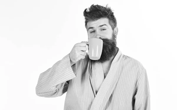 Человек с бородой и усами держит кружку кофе, изолированного на белом. Утренний кофе. Мачо в халате пьет чай или кофе со спящим лицом. Парень выглядит сонным, ему нужен кофе. — стоковое фото
