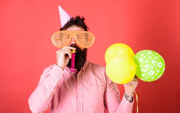 Mann mit Bart auf ruhigem Gesicht mit Partyhorn und Luftballons, roter Hintergrund. Mann im Partyhut mit Feiertagsattributen feiert. Überraschungskonzept. Hipster mit Riesenbrille bläst in Partyhorn — Stockfoto