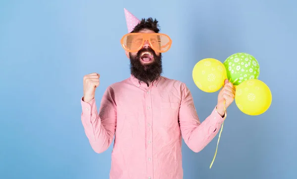 Hipster mit Riesenbrille feiert Geburtstag. Festkonzept. Mann mit Bart auf fröhlichem Gesicht hält Luftballons, hellblauer Hintergrund. Mann mit Partyhut und Luftballons feiert — Stockfoto