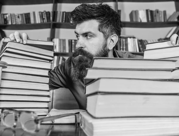 Profesor o estudiante con barba se sienta en la mesa con gafas, desenfocado. Concepto bibliófilo. Hombre en cara estricta entre montones de libros, mientras estudia en la biblioteca, estanterías en el fondo —  Fotos de Stock