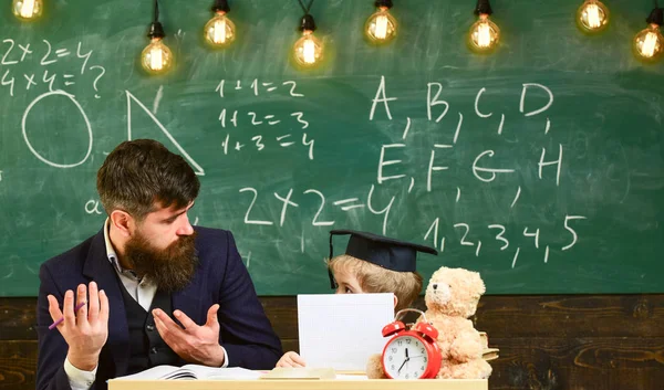 Grade school concept. Teacher and pupil in mortarboard, chalkboard on background. Kid studies with teacher, listening with attention. Father teaches son elementary knowledge, discuss, explain
