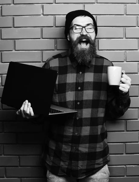 Bearded brutal caucasian hipster holding laptop with cup — Stock Photo, Image