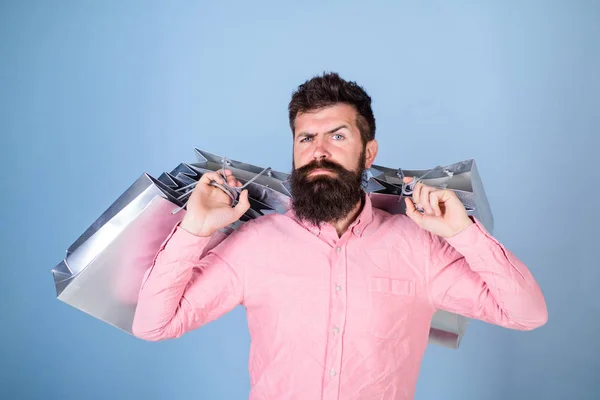 Concepto de venta y descuento. Hipster en la cara estricta de compras adicto o adicto a las compras. Hombre con barba y bigote llevar bolsas de compras, fondo azul claro. Novio llevando un montón de bolsas de compras —  Fotos de Stock