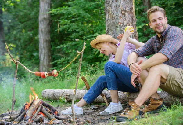 Have snack. Picnic roasting food over fire. Cooking at picnic. Girl offers eat apple while they waiting roasted food. Family enjoy romantic weekend in nature. Couple relaxing sit on log having snacks