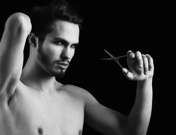 Schöner bärtiger Macho mit stylischem Haar auf ernstem Gesicht und nackter Brust mit Schere im Studio auf schwarzem Hintergrund. — Stockfoto