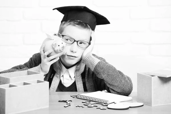 Young cute pupil boy in grey sweater and glasses sitting at desk learned on hand with copybook wooden numbers pink piggy pig bank in graduation cap in classroom on white brick wall background — Stock Photo, Image