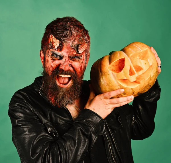 Man wearing scary makeup holds pumpkin on green background.