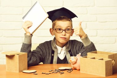 Gri süveterli, gözlüklü, elinde defteriyle masada oturan yakışıklı öğrenci. Ahşap numaralar. Mezuniyet şapkasında pembe domuzcuk bankası.