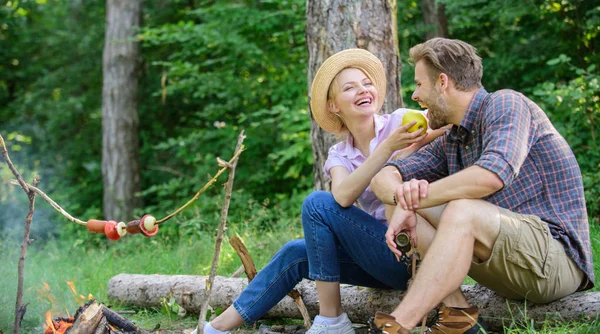 Have snack. Couple relaxing sit on log having snacks. Picnic roasting food over fire. Cooking at picnic. Girl offers eat apple while they waiting roasted food. Family enjoy romantic weekend in nature