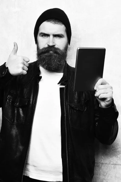 Pria berjenggot, jenggot panjang. Kaukasia brutal hipster yang tidak dicukur serius memegang tablet dengan jaket kulit hitam dengan latar belakang dinding tekstur yang babak belur. — Stok Foto