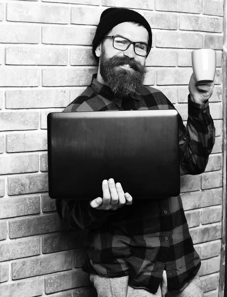 checkered shirt with hat and glasses on beige brick wall studio background. Bearded brutal caucasian hipster holding laptop with cup