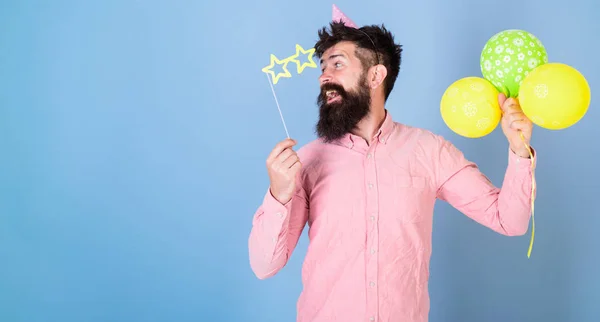 Homem com barba no rosto surpreso segurar balões de ar, fundo azul claro. Hipster em óculos em forma de estrela celebra o aniversário. Partido atribui conceito. Cara em chapéu de festa com balões de ar celebra — Fotografia de Stock