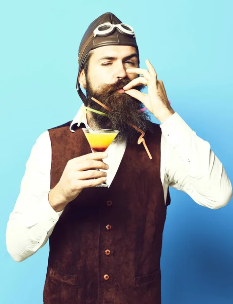 Homem aviador barbudo bonito com barba longa e bigode no rosto satisfeito segurando vidro de bebida alcoólica em jaqueta de couro de camurça vintage com chapéu e óculos no fundo do estúdio azul — Fotografia de Stock