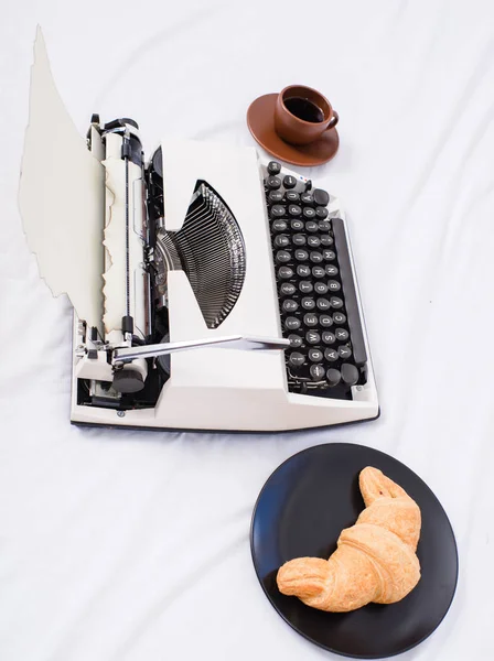 Benefit Schriftsteller zu sein ist ein angenehmer inspirierender Arbeitsplatz. schaffen eine inspirierende Atmosphäre, bevor Sie mit dem Schreiben einer Schreibmaschine beginnen. Schreibmaschine und köstliche Croissanteller und Kaffee weiße Bettwäsche — Stockfoto
