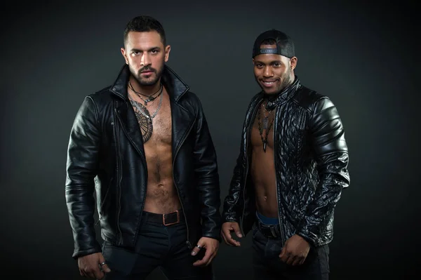 Machos with muscular torsos look attractive in leather jackets, dark background. Men on confident faces with bristle. Men with sexy muscular torsos look brutally. Masculinity and brutality concept