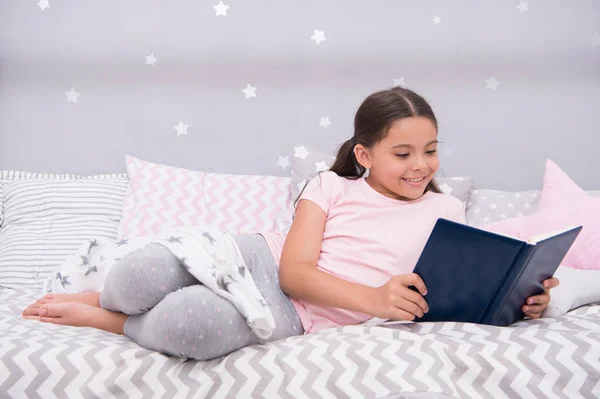 Girl child lay bed read book. Kid prepare to go to bed. Pleasant time in cozy bedroom. Girl kid long hair cute pajamas relax and read book. Satisfied with happy end. She likes kind stories about love