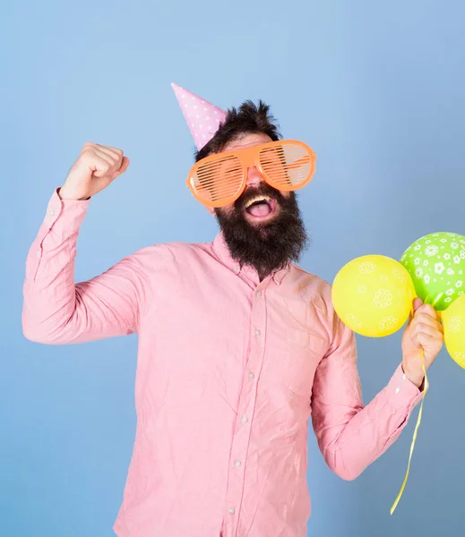Hipster in giant eyeglasses celebrates birthday. Guy in party hat with air balloons celebrates. Celebration concept. Man with beard on cheerful face hold air balloons, light blue background — Stock Photo, Image