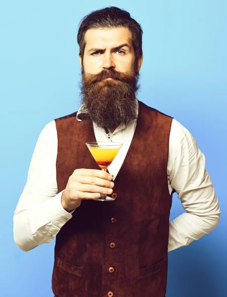 Handsome bearded man with long beard and mustache has stylish hair on serious face holding glass of alcoholic cocktail in vintage suede leather waistcoat on blue studio background — Stock Photo, Image