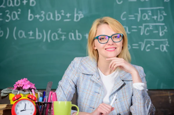 Arbeitsbedingungen, die angehende Lehrer berücksichtigen müssen. Arbeitsbedingungen für Lehrer. Arbeit weit über den eigentlichen Schultag hinaus. Frau lächelnd Lehrer sitzen Tisch Klassenzimmer Kreidetafel Hintergrund — Stockfoto