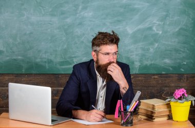 Evrak öğretmenler hayatın bir parçası. Ödev kontrol edin. Öğretmen hipster gözlük sit sınıf kara tahta arka plan ile sakallı. Ev ödevleri veya test kontrol öğretmen. Öğretmen Danışma dizüstü bilgisayar ile oturup