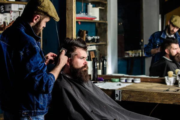 Hipster client getting haircut. Barber with hair clipper works on hairstyle for man with beard, barbershop background. Barber styling hair of bearded client with comb and clipper. Haircut concept