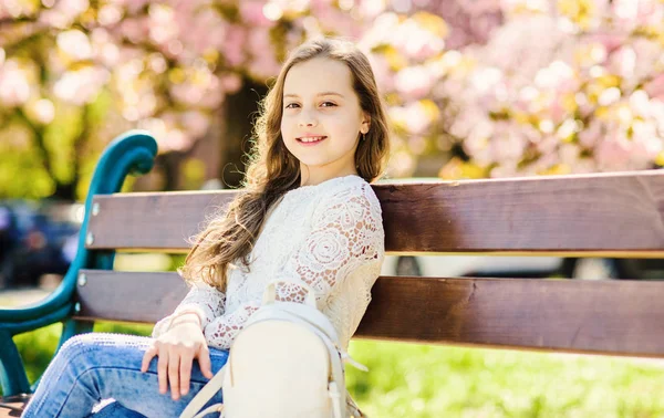 Lány a mosolygó arc ül a padon, sakura fa a háttérben, defocused. Hosszú gyönyörű haj aranyos gyermek élvezze a napos Tavasz. Lány pihentető Park közelében cherry blossom. Természetes szépség fogalma — Stock Fotó