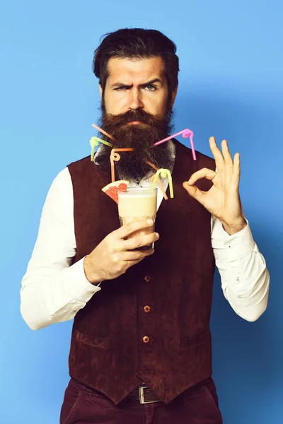 Homem barbudo bonito com tubos coloridos em barba longa e bigode tem cabelo elegante no rosto sério segurando vidro de coquetel alcoólico na cintura de couro de camurça vintage no fundo do estúdio azul — Fotografia de Stock