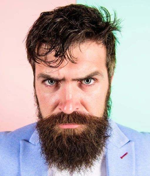 Man bearded hipster on strict face pink blue background. Hipster guy with messy tousled hair and long beard needs barber service. Keep hair tidy and care about hairstyle. Barber tips grooming beard.