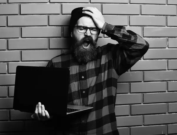 Homme barbu, longue barbe. Brutal caucasien bâillement hipster non rasé tenant ordinateur portable en rouge noir chemise à carreaux avec chapeau et lunettes sur fond de studio mur de brique marron . — Photo