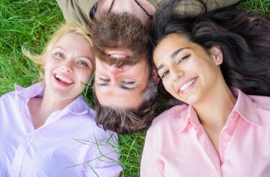 Blonde and brunette lay on his shoulders. Man fall in love both women. Hipster relaxing pleasant women company. Happy threesome. Man bearded hipster lay on grass with two girls blonde and brunette clipart