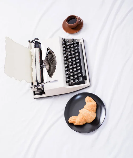 Schreibmaschine und köstliche Croissanteller und Kaffee weiße Bettwäsche. schaffen eine inspirierende Atmosphäre, bevor Sie mit dem Schreiben einer Schreibmaschine beginnen. Schriftsteller zu sein ist ein angenehmer inspirierender Arbeitsplatz — Stockfoto