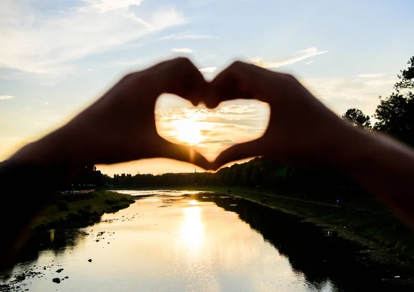 Heart gesture in front of sunset above river. Idea honeymoon travel. Honeymoon summer resort. Romantic date ideas. Hands heart gesture symbol of love and romance. Sunset sunlight romantic atmosphere