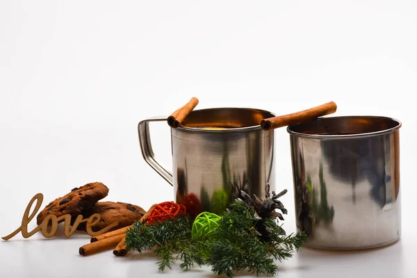 Mugs with mulled wine or hot drink near winter decoration and cookies on white background, close up. Mulled wine or hot beverage in metal mugs with cinnamon sticks and word love. Hot wine concept