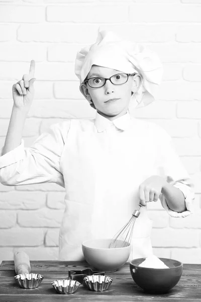Мальчик маленький милый повар-повар в белой униформе и шляпе на удивленном лице с очками, стоящими рядом со столом с роликовой формы булавки для тортов и приготовления пищи на фоне кирпичной стены — стоковое фото