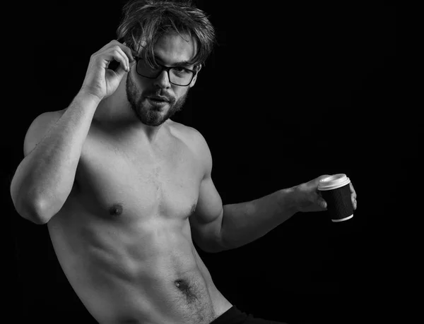 handsome bearded athletic macho man with stylish beard standing in glasses with muscle torso on athletic body holding hot coffe cup in studio on black background