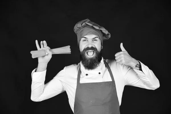 Koken met vrolijk gezicht in Bourgondië hoed en schort houdt droge pasta. Chef-kok met bos van spaghetti verschijnt duim — Stockfoto