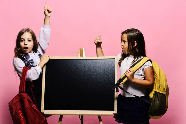 Las chicas con caras sorprendidas sostienen los dedos para arriba teniendo idea. Los niños que usan bolsos de escuela se apoyan en la pizarra, espacio para copiar . —  Fotos de Stock