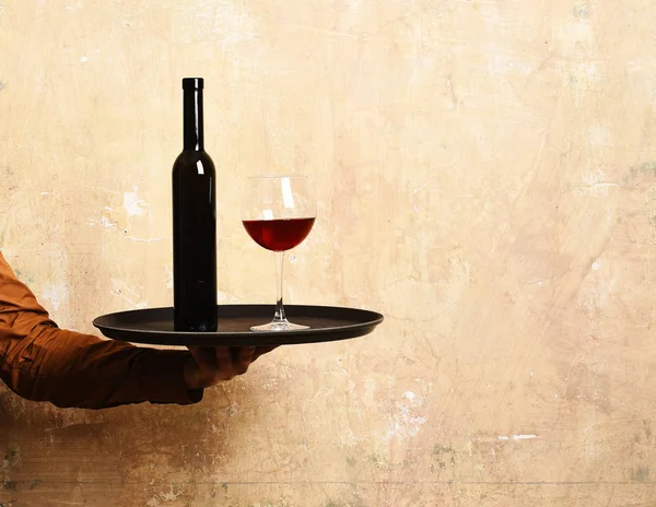 Waiter with glass and bottle of red wine on tray. Barmans hand holds and serves red wine. Service and catering concept. Male hand holds wine on beige wall background, copy space.