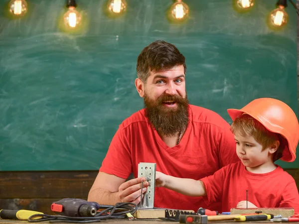 Father with beard teaching little son to use tools in classroom, chalkboard on background. Boy, child in protective helmet makes by hand, repairing with cheerful dad. Little assistant concept